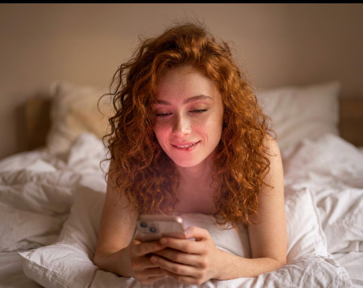 A woman lies in bed smiling softly at her phone, warm light on her face.